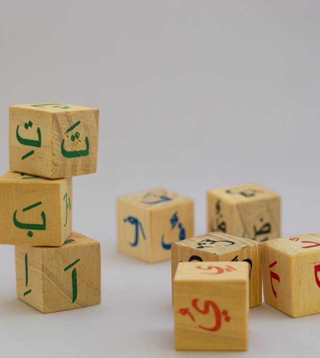 Tower of wooden blocks with green Arabic letters, set on a pale surface with other blocks nearby.