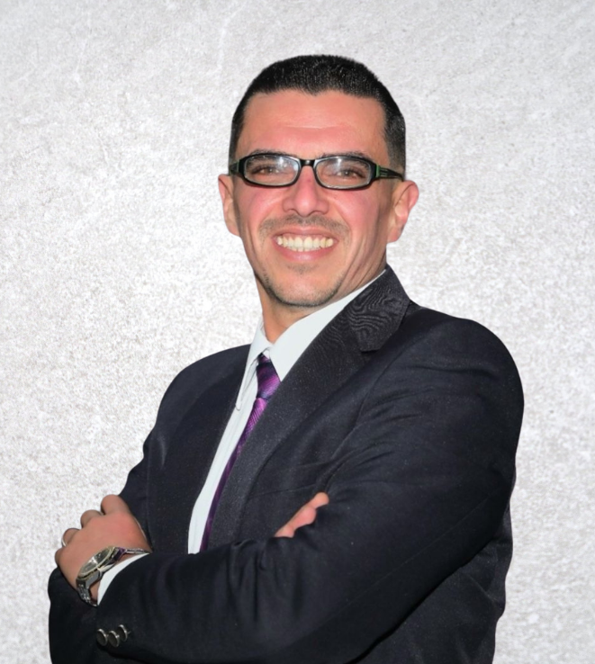 Man in a dark suit and purple tie, smiling with arms crossed against a light gray textured background.